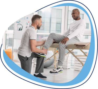A healthcare professional assists a patient with a leg stretch in a therapy room. The patient, sitting on a treatment table, is receiving care for his leg while the therapist gently supports his limb. The setting is bright and modern.